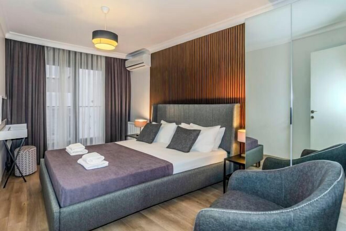 A modern hotel room with a large bed, bedside tables, a chair, a desk, and a wardrobe.