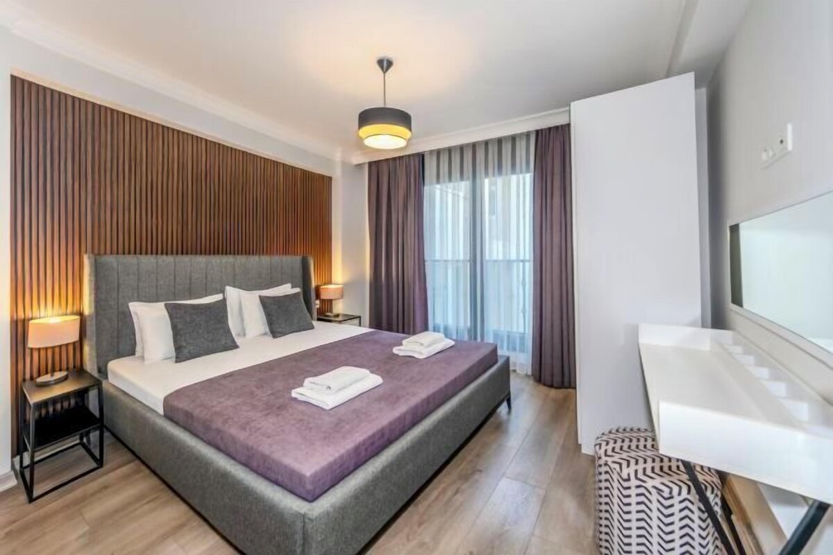 A modern hotel room with a large bed, bedside lamps, a flat-screen TV, and a bathroom with a white sink.