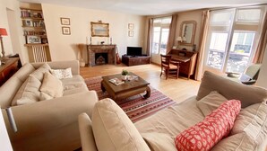 A living room with a fireplace, a television, a dining area, and a large window with a balcony. - RIVE GAUCHE - RIVOLI/LOUVRE Type 3/4 approx. 100m2 in absolute calm (Paris)
