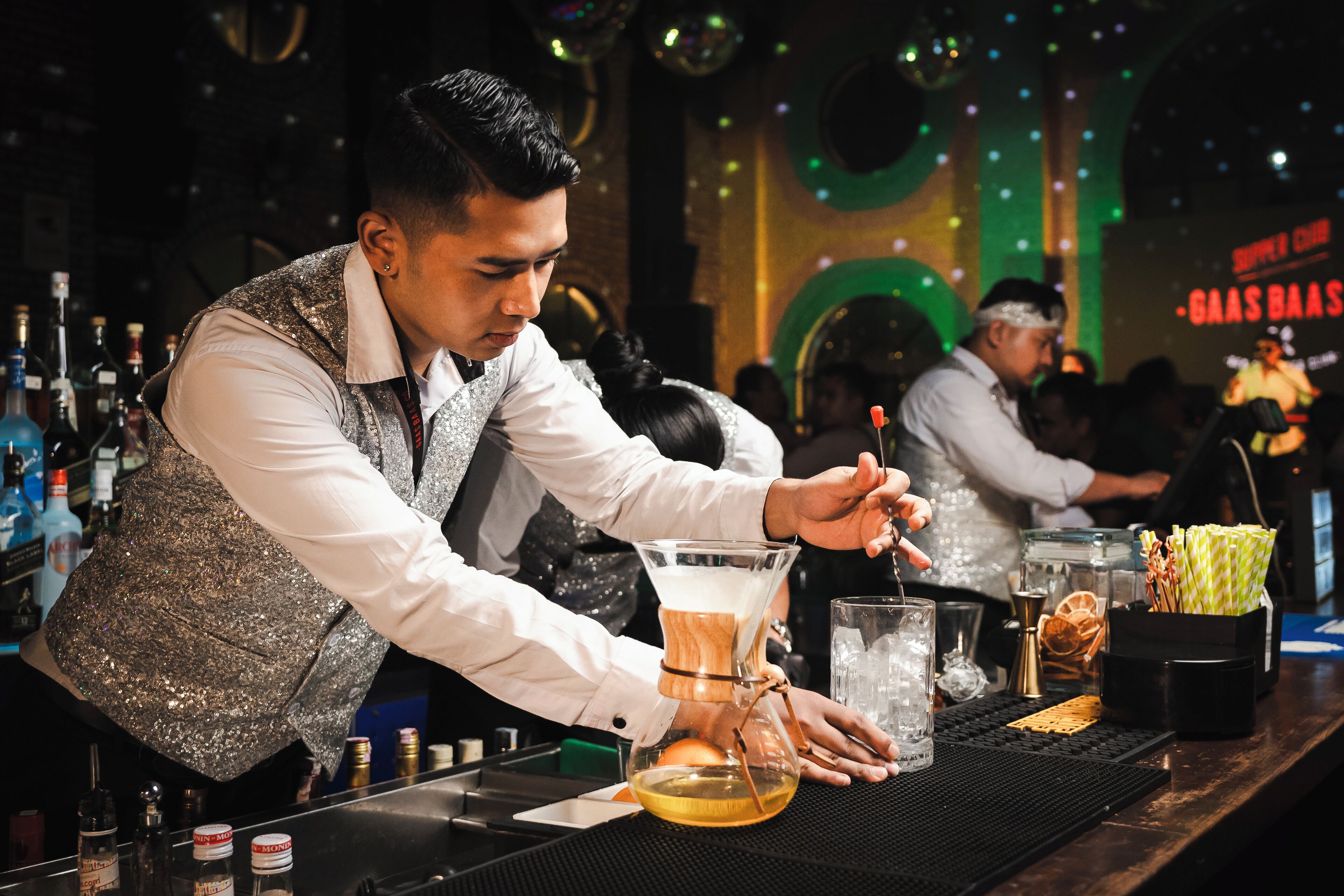 A bartender preparing a drink at a bar with a variety of bottles and a glass on the counter.