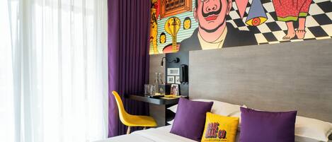 A hotel room with a bed, a desk, and a vibrant mural on the wall.