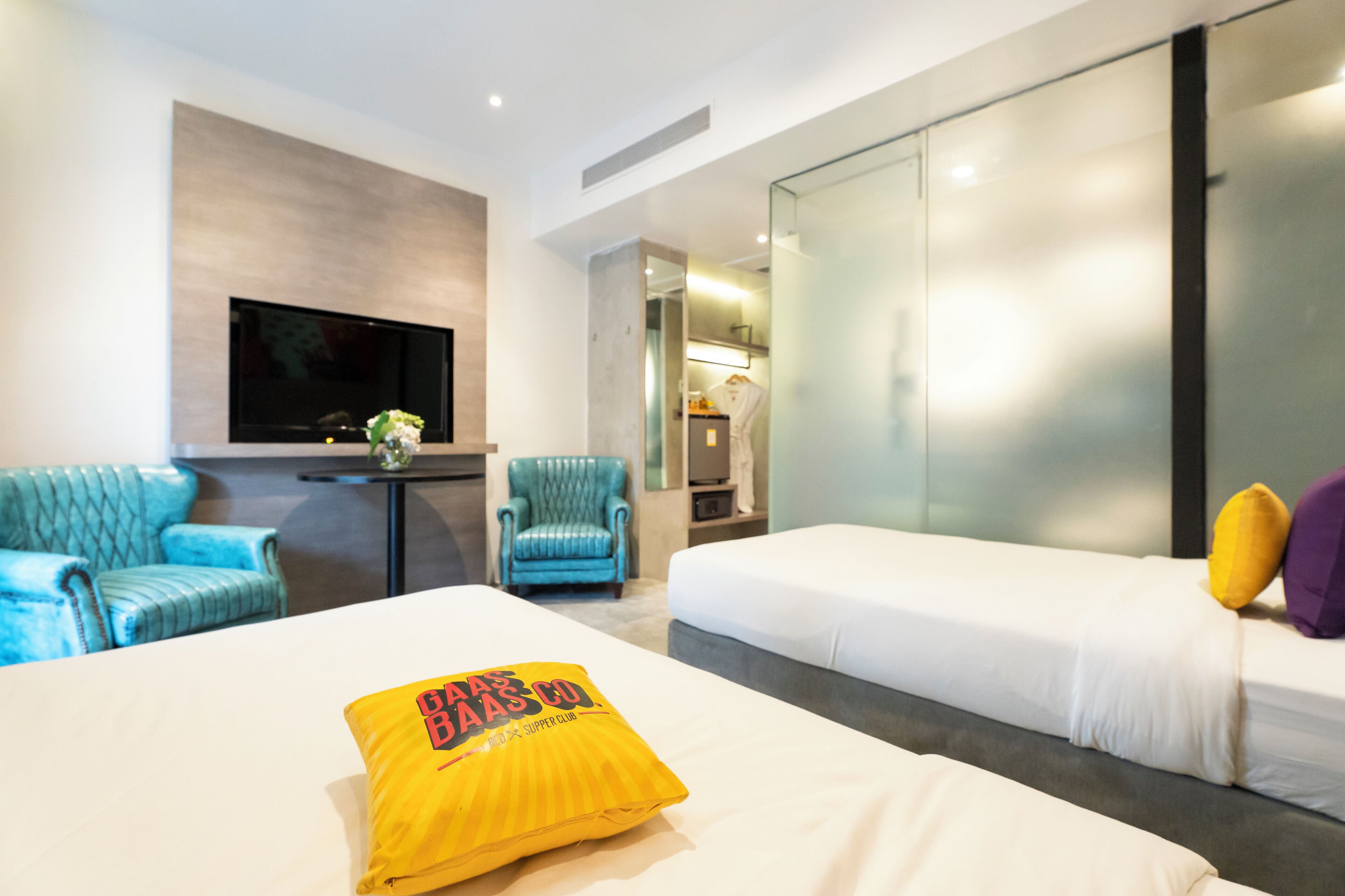 A modern hotel room with a large bed, a TV mounted on the wall, a seating area with blue armchairs, and a glass partition separating the room from the bathroom.
