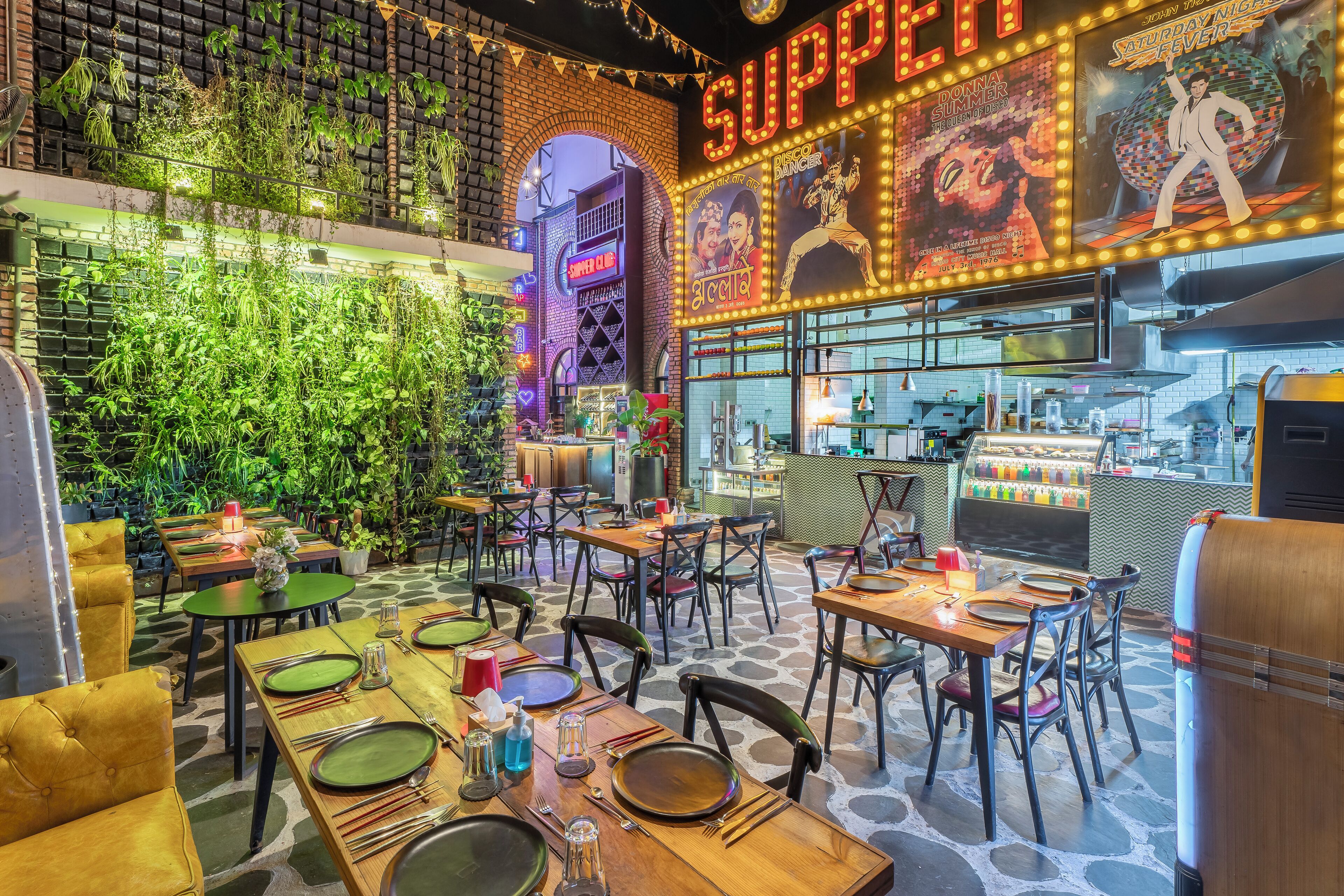 An indoor dining area with tables set for multiple people, a brick wall with greenery, and a neon sign advertising 'SUPPER'.