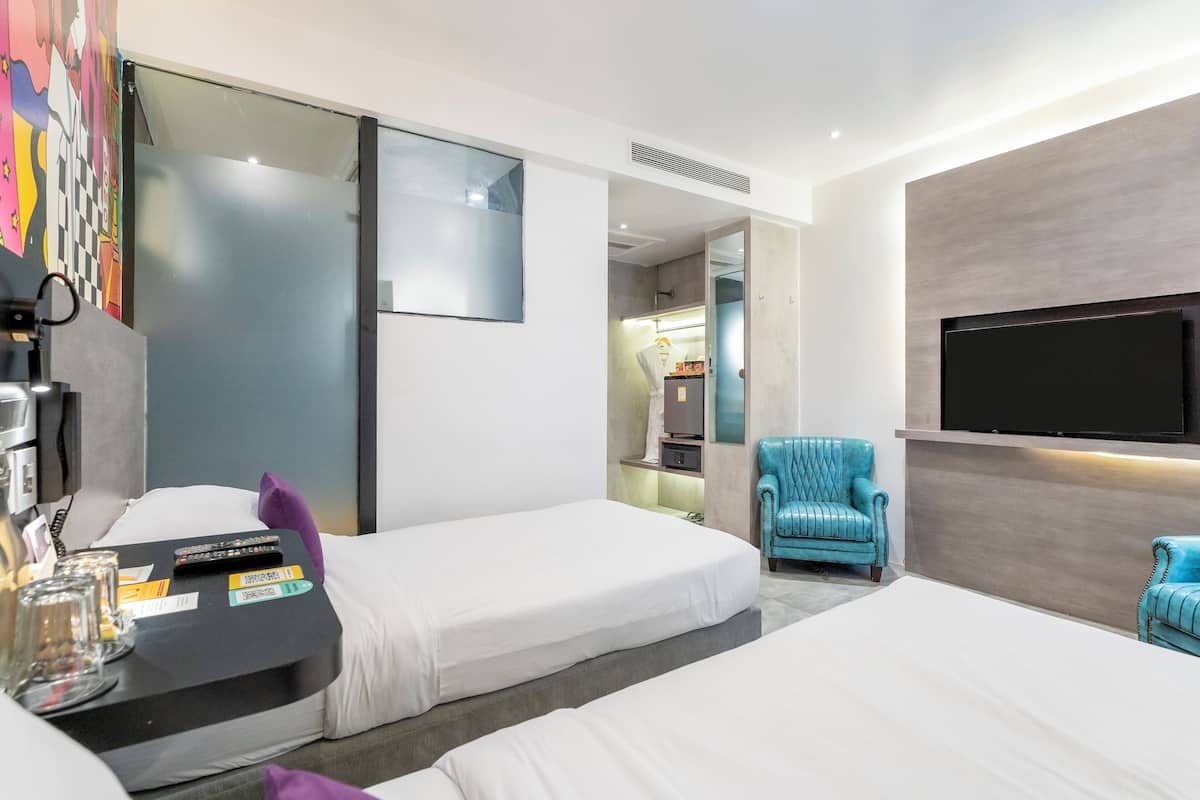Ein Hotelzimmer mit zwei Einzelbetten, einem an der Wand befestigten Fernseher, einem kleinen Schreibtisch mit Lampe und einem blauen Sessel.