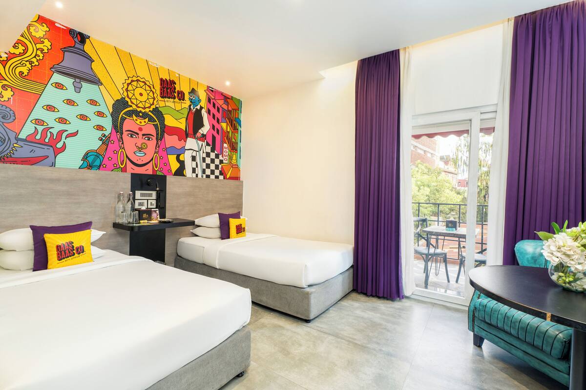 Ein Hotelzimmer mit zwei Betten, einem farbenfrohen Wandgemälde, einem Balkon mit violetten Vorhängen und einer Essecke mit einer senffarbenen Bank.