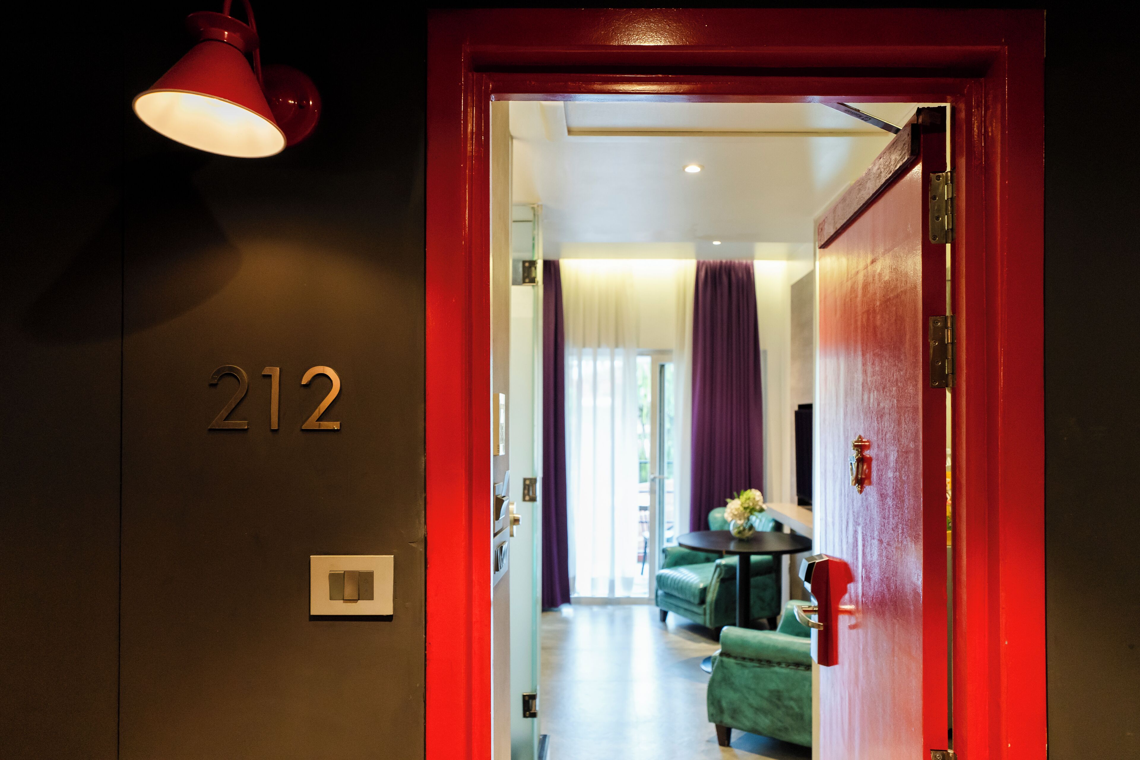 A hotel room entrance with a red door, a number '212' on the wall, a light fixture, and a glimpse of a green sofa and curtains inside.