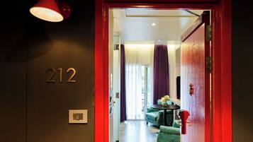 A hotel room entrance with a red door, a number '212' on the wall, a light fixture, and a glimpse of a green sofa and curtains inside.