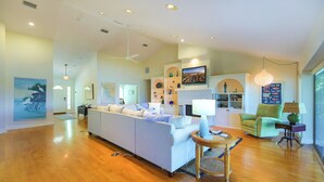 A spacious living room with a large sectional sofa, a dining area with a table and chairs, and a kitchen area with a TV mounted above the counter. - Sanibel Sunrise - Bayfront home, short walk to the beach! (Sanibel)