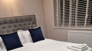 A neatly made bed with white linens and folded towels, a tufted headboard, and a window with blinds. - Beautiful and Modern House in Gravesend (Gravesend)