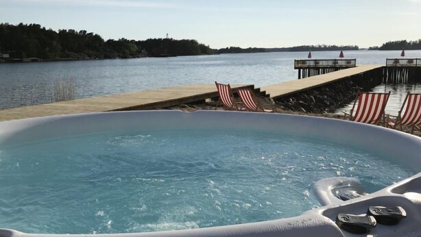 A hot tub with a wooden deck and American flags, overlooking a body of water and a dock. - Carlsro Badhotell (Föglö)