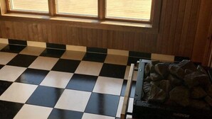 A room with a large window overlooking a wooden deck and a body of water. There is a checkered floor and a Harvia fireplace. - Carlsro Badhotell (Föglö)
