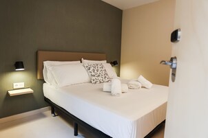 Una cama bien hecha con sábanas blancas y un cojín floral, una mesita de noche con lámpara, y un enchufe en la pared.