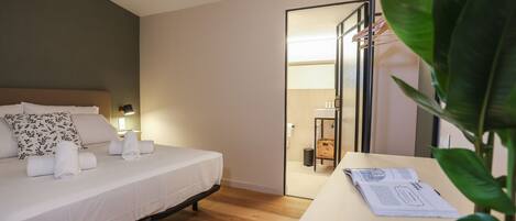 A neatly made bed with white linens and a floral pillow, a bedside table with a book and a potted plant, and a wooden floor.
