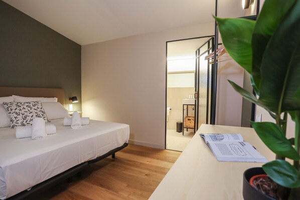 Una cama bien hecha con sábanas blancas y una almohada con motivos florales, una mesita de noche con un libro y una planta en maceta, y un suelo de madera.