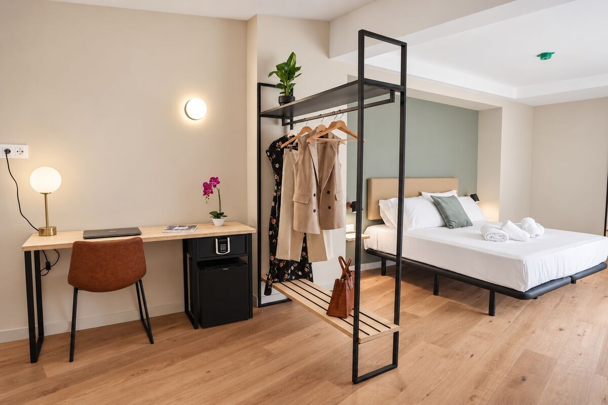 A modern hotel room with a desk, chair, bed, and a clothing rack.