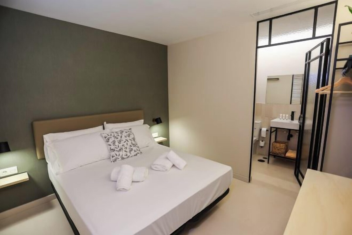 A hotel room with a bed, a nightstand, a mirror, and a bathroom visible through a glass door.