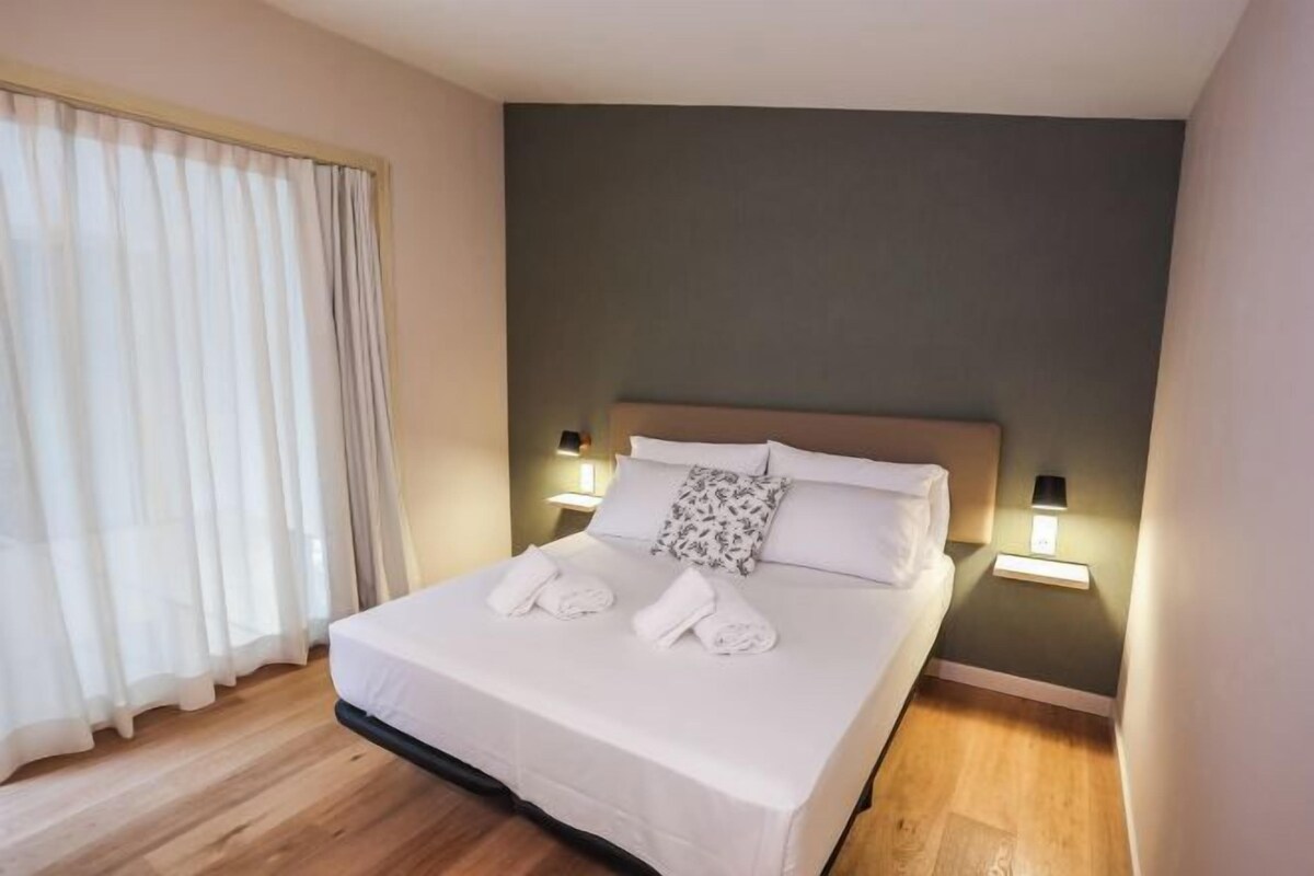 A neatly made bed with white linens and towels, a wooden floor, and two bedside lamps.