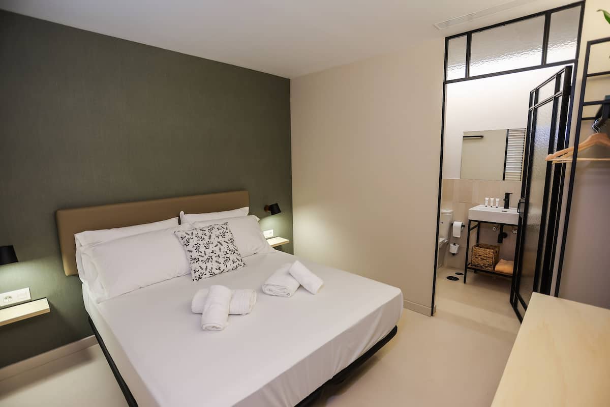 A neatly made bed with white linens and two rolled towels, a bedside table with a lamp, and a small bedside shelf.