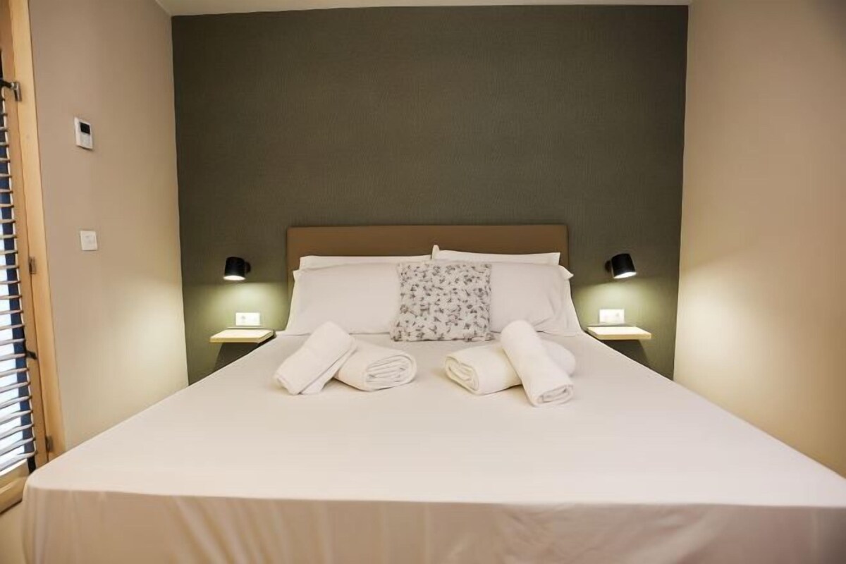 Una cama bien tendida con sábanas blancas y un cojín floral, dos lámparas de noche y una mesita con lámpara.