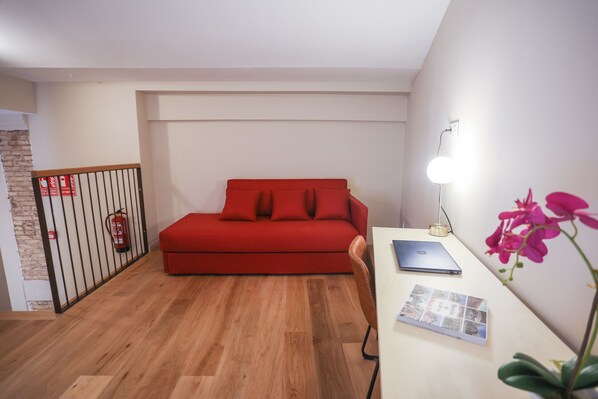 A room with a red sofa, a wooden floor, a desk with a laptop, a lamp, and a plant. - Mythic Valencia Hotel (Valencia)