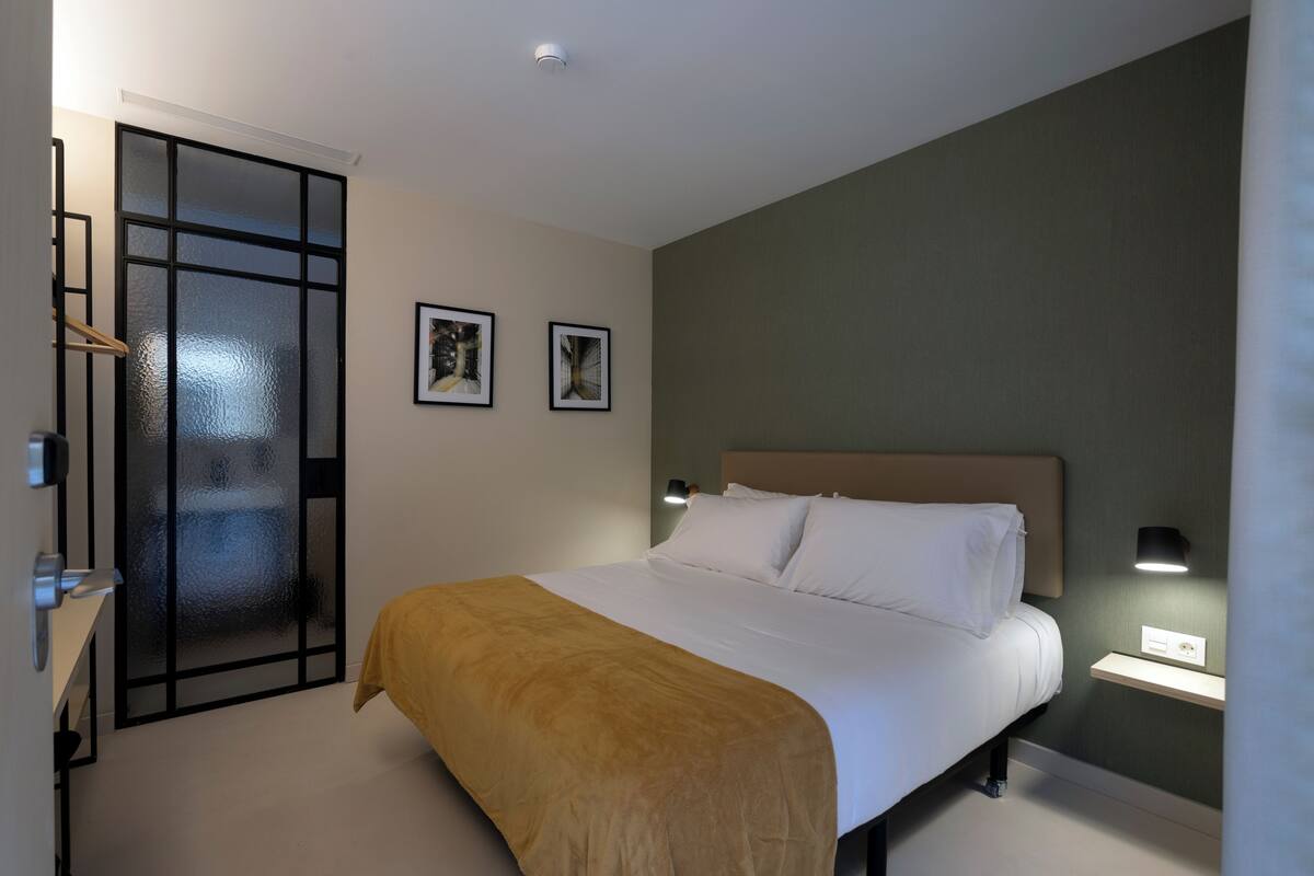 A modern hotel room with a large bed, a bedside table, a wall-mounted light, and a door with frosted glass panels.