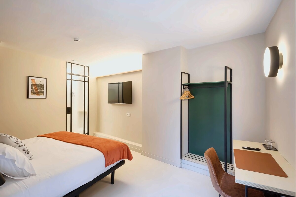 A modern hotel room with a bed, a desk, a chair, a wardrobe, and a mirror.