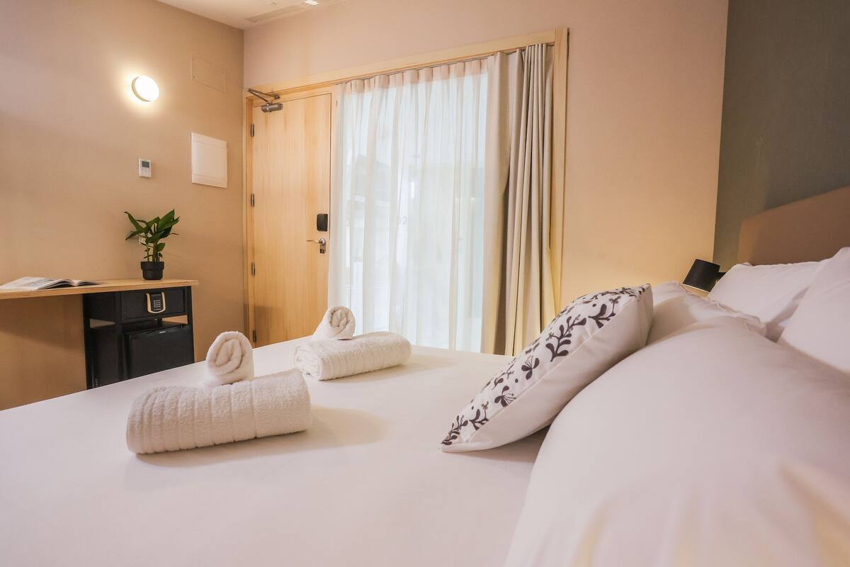 A hotel room with a bed, two towels, a desk, a plant, and a window with curtains.