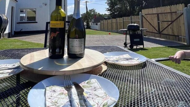 Eine Terrasse mit rundem Tisch, gedeckt für zwei Personen, mit einer Weinflasche und einem Geschirr mit Besteck.