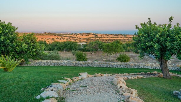 A landscaped garden with a stone pathway, trees, and a view of a distant landscape. - Rosemary - open space in the Hyblean countryside (Sicilia)