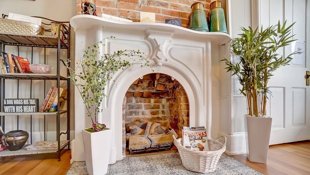 A fireplace with a brick interior, surrounded by a white mantelpiece. A bookshelf with various books and decorative items is to the left, and a potted plant is to the right. - Modern Rustic Charm with City Views (Cincinnati)