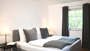 Una cama bien hecha con ropa de cama blanca y gris, una lámpara de noche y una ventana con cortinas.