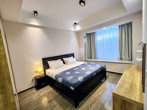 Hotellihuone, jossa on sänky, yöpöytä, lamppu ja ikkuna, jossa on verhot.