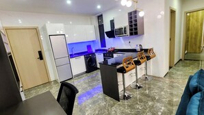 Een moderne keuken met een ingebouwde oven, magnetron en spoelbak. Drie barstoelen en een eettafel zijn aanwezig.