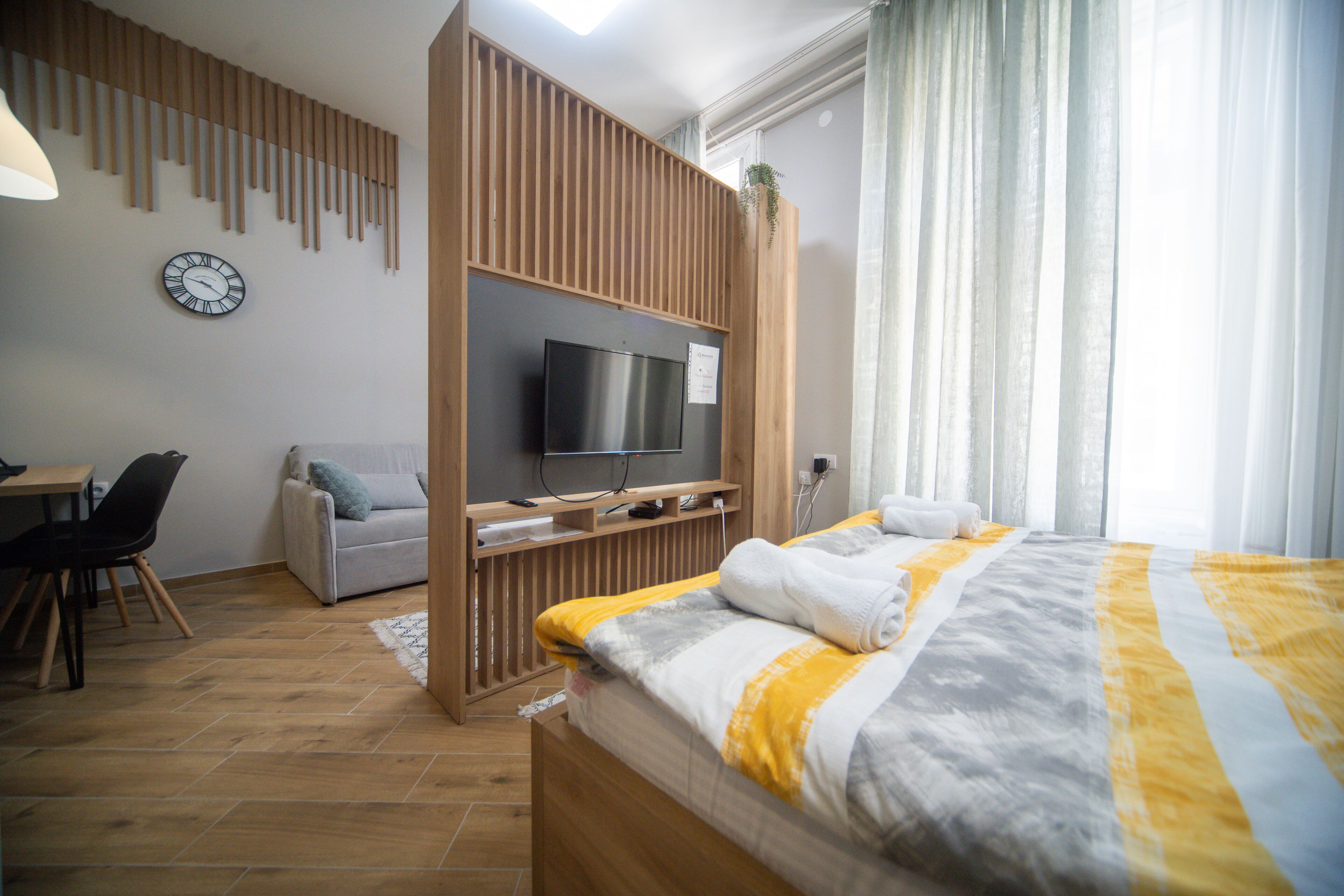 A modern hotel room with a wooden TV stand, a bed with a striped blanket, a desk with a chair, and a large window with curtains.