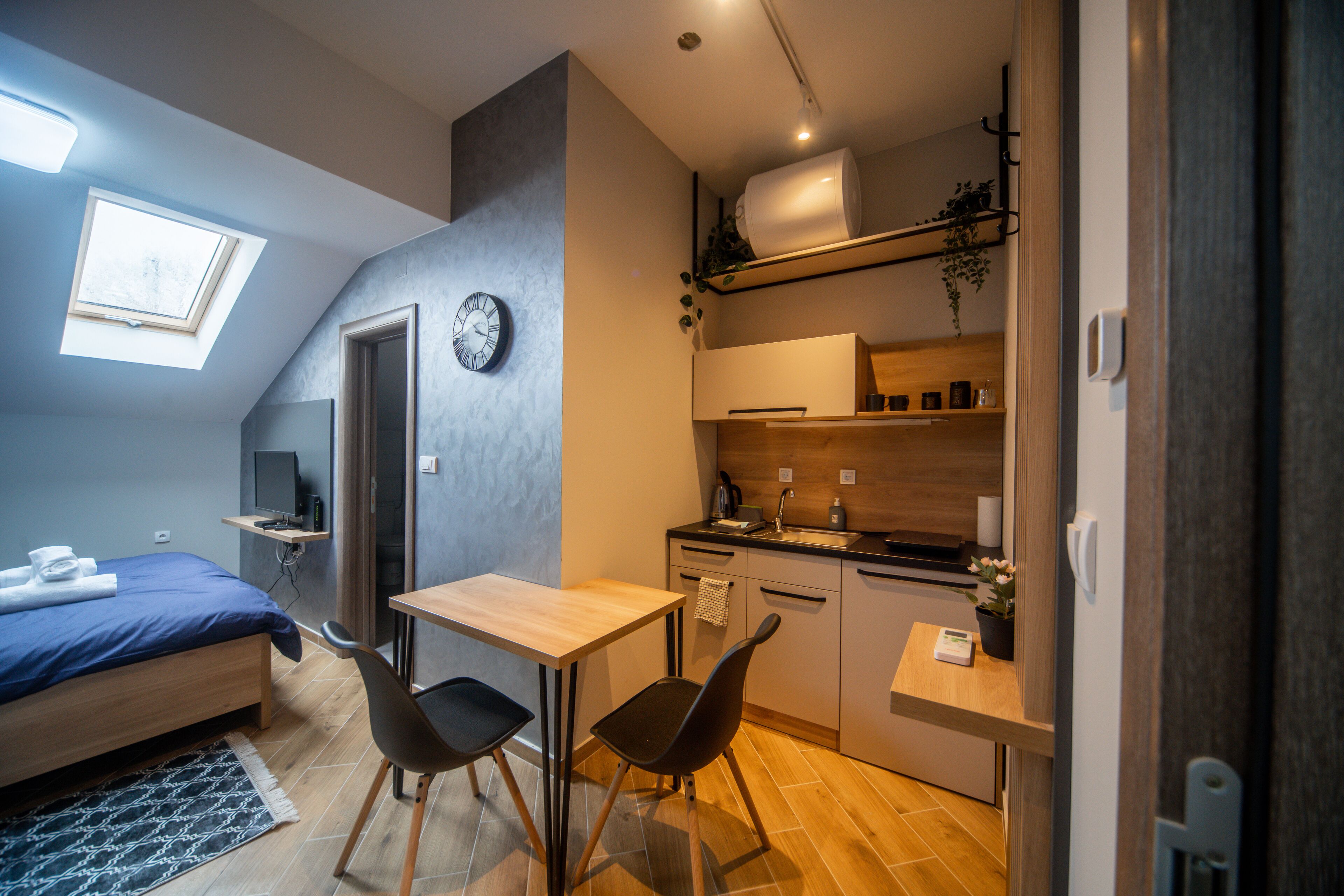 A compact bedroom with a bed, bedside table, and a small desk with a chair. A skylight provides natural light, and a clock is visible on the wall.
