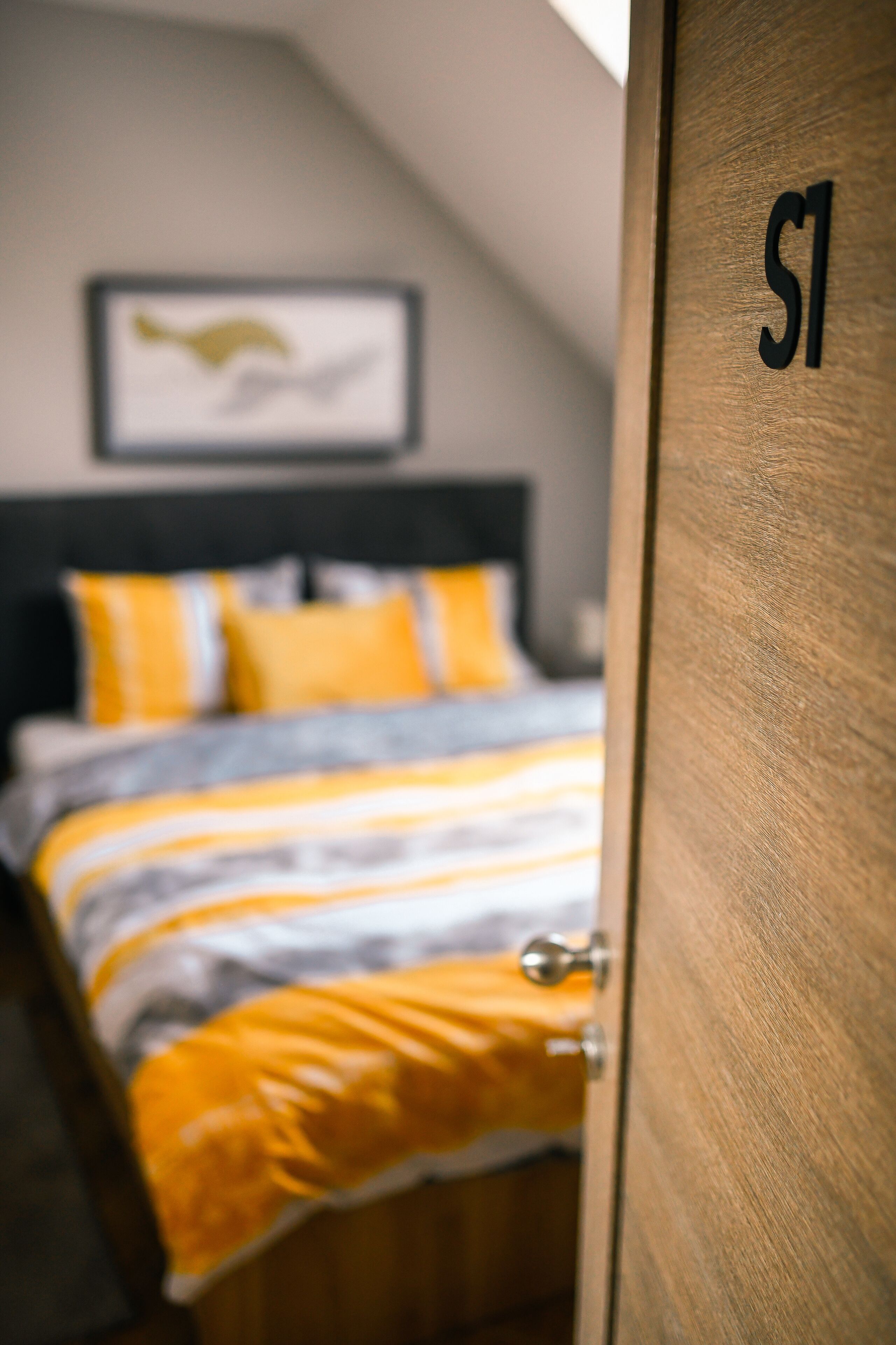 A bedroom with a bed, a wooden door with a number, and a framed picture on the wall.