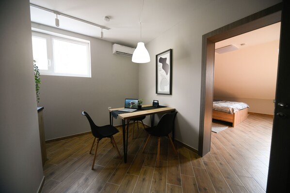 Une pièce de taille modeste avec un plancher en bois, un bureau avec un ordinateur portable, deux chaises noires, un luminaire suspendu, un tableau encadré et un lit placé dans un coin.