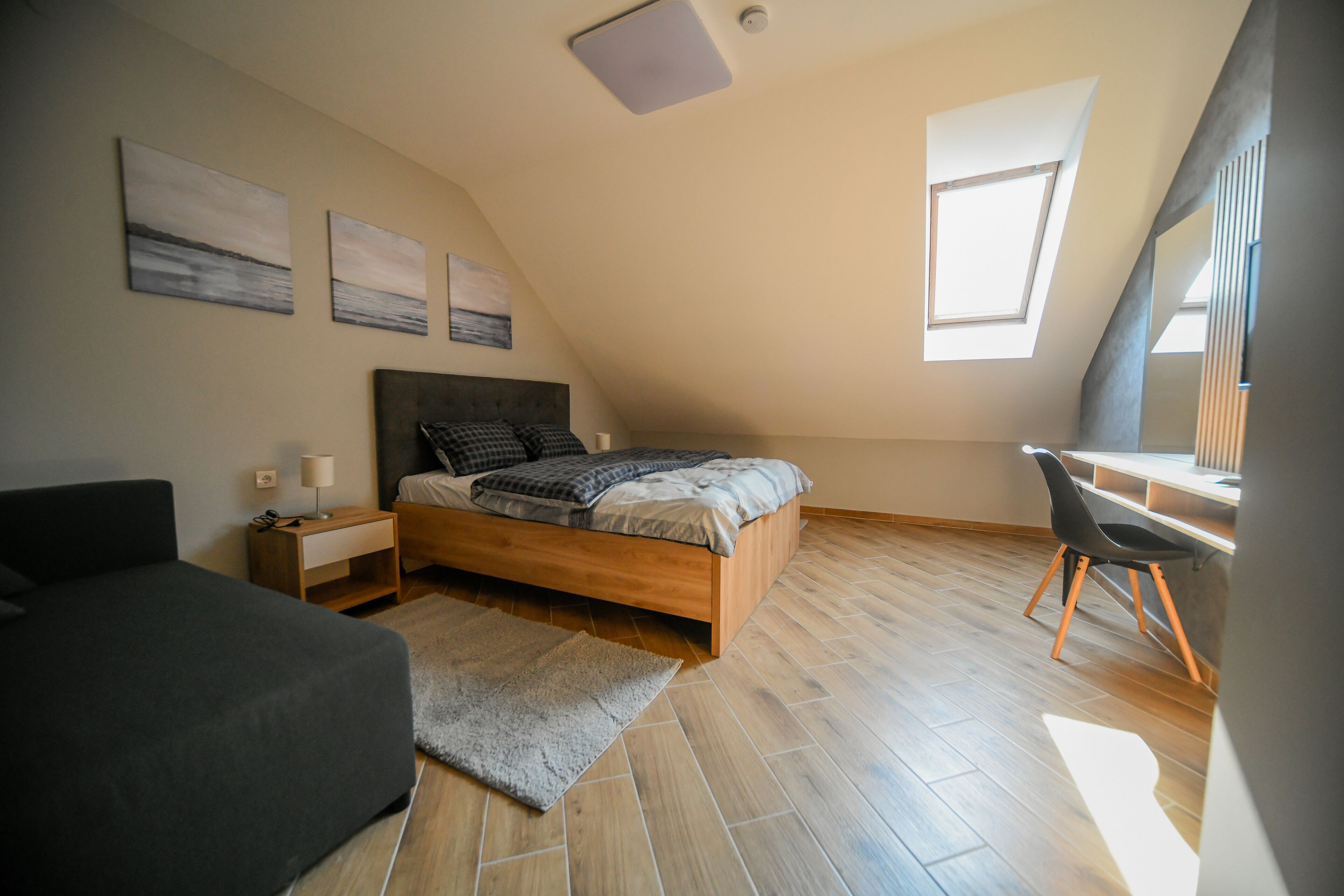 A bedroom with a bed, bedside table, lamp, and a desk with a chair. There are three paintings on the wall and a skylight.