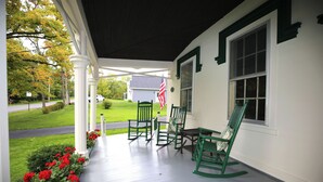 A porch with white columns, green rocking chairs, and a view of a house with an American flag. - Historic Home at Sheldrake in the Finger Lakes. (Ovid)