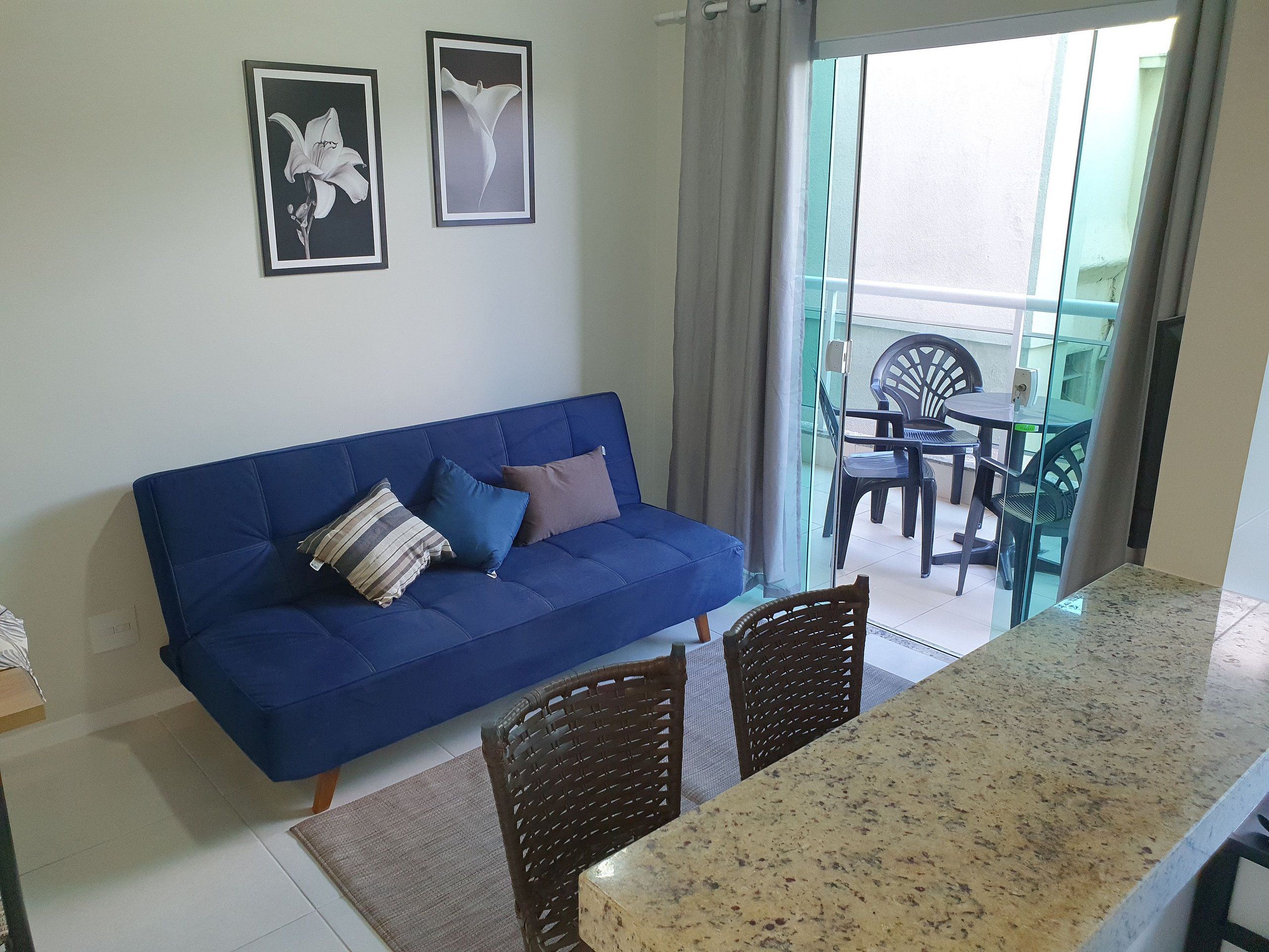 A blue sofa with cushions, a dining table with chairs, and a balcony with plastic chairs and a table.
