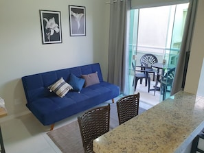 A blue sofa with cushions, a dining table with chairs, and a balcony with plastic chairs and a table. - AP encantador na Praia do Forte! (208) (Cabo Frio)