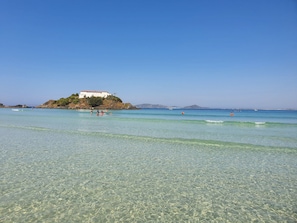 A beach with clear water, a white building on a small island, and a few people in the water. - AP encantador na Praia do Forte! (208) (Cabo Frio)
