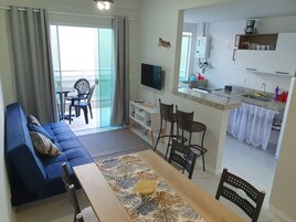 Uma sala de estar com sofá, uma mesa de jantar com jogo americano floral, televisão e uma cozinha com cafeteira e armários.