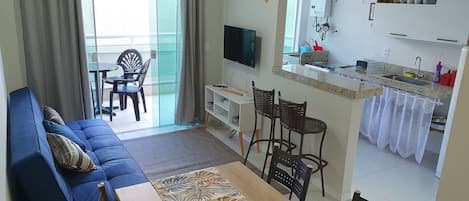 A living room with a sofa, a dining table with chairs, a TV, and a kitchen area with a sink and cabinets.