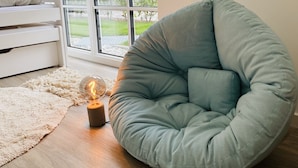 Une pièce avec une grande fenêtre, un fauteuil en forme de poire de couleur bleu clair, une petite table avec une lampe en verre et un canapé blanc.