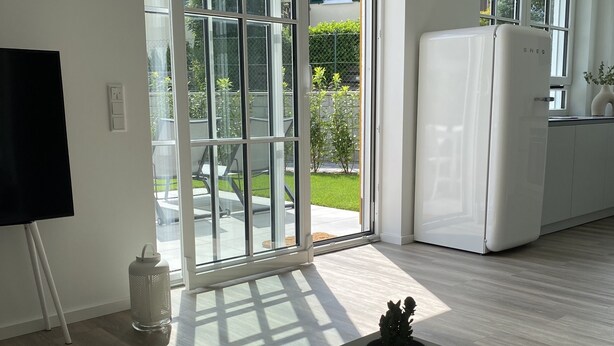 Une cuisine moderne avec un réfrigérateur blanc, un coin repas doté d’une table blanche et une porte-fenêtre coulissante donnant sur un jardin.