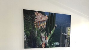 Une chambre à coucher comprenant un lit, deux tables de chevet, une lampe fixée au mur et un grand tableau représentant une maison et des arbres.