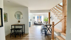 Un intérieur moderne avec un escalier en bois, un coin repas avec des tabourets, un canapé et une cuisine en arrière-plan.