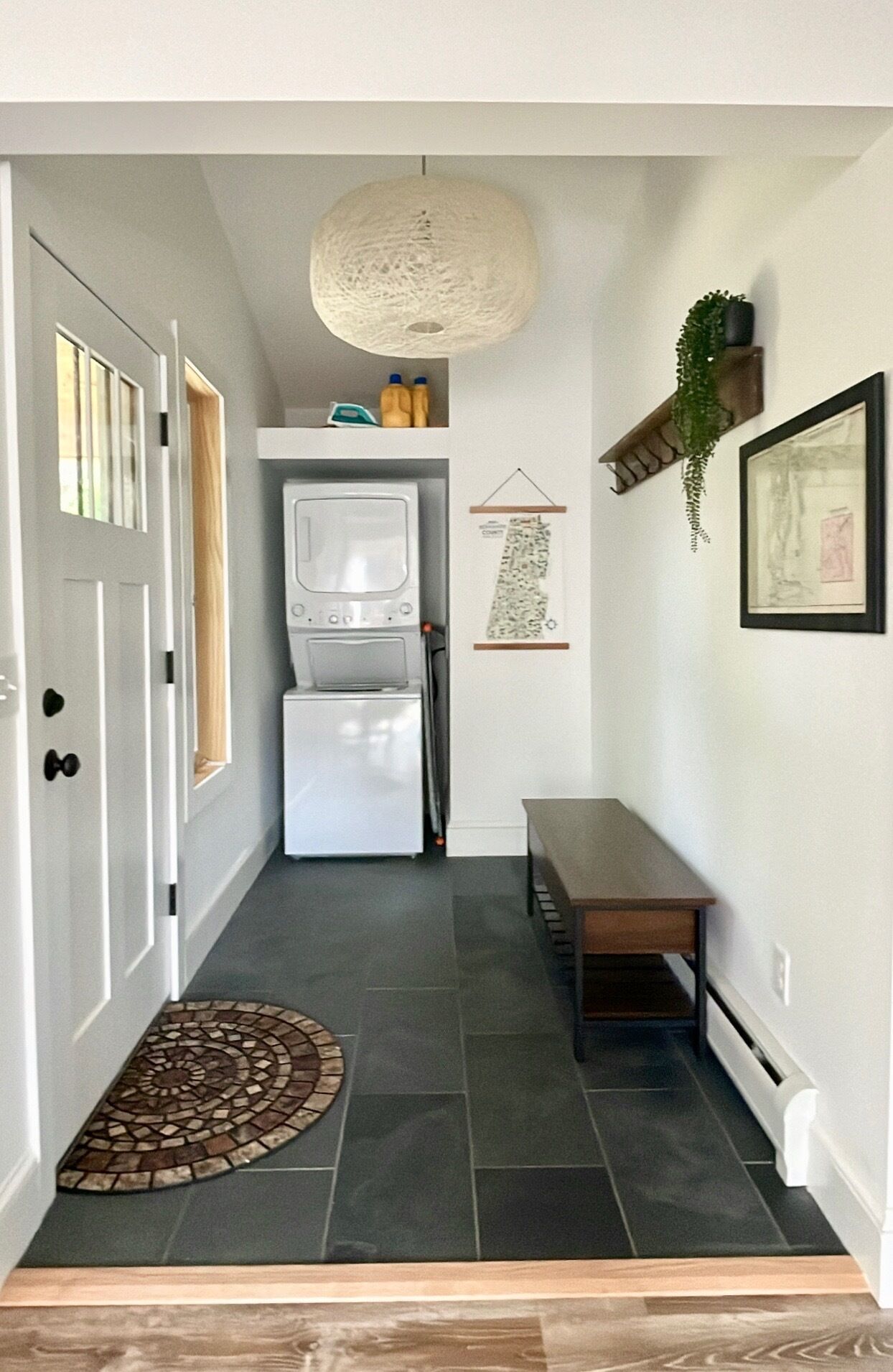 A narrow hallway with a white door, a white refrigerator, a white oven, a bench, a hanging light fixture, and a small shelf with a plant.