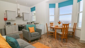A modern living room with a grey sofa, orange throw, and teal pillows. A dining area with wooden chairs and a table is adjacent to a kitchen with white cabinets and a microwave. Large windows with blue blinds offer a view of the sea. - Apartment 6 Bay View (Scarborough)